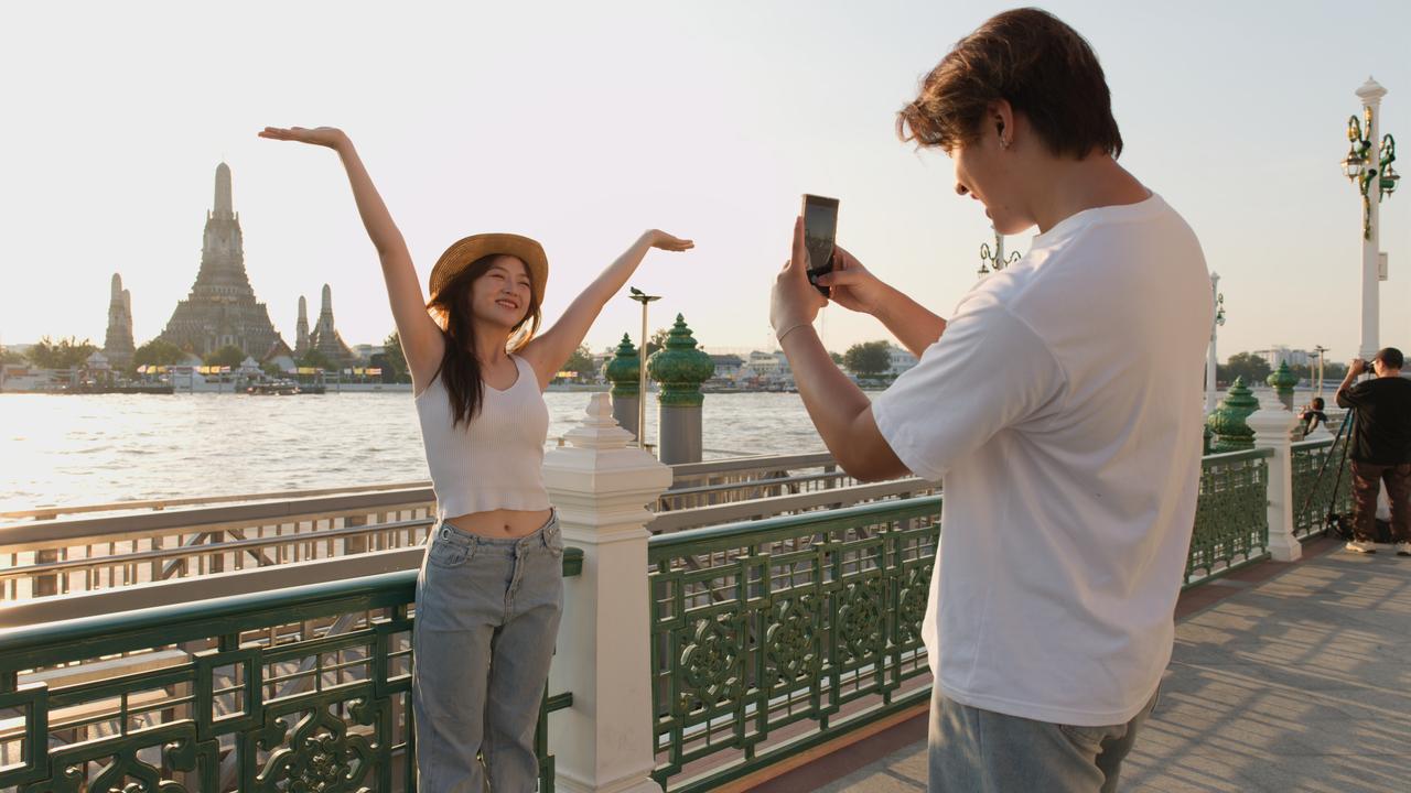Young Couple Taking Photos by Chao Phraya River with Wat Arun Background. (Adobe Stock photo)