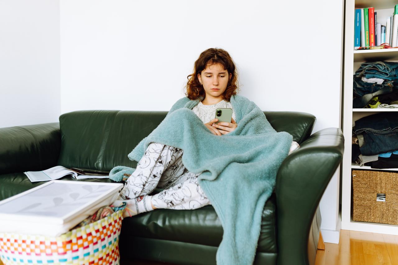 A young girl sits on a sofa wrapped in a blanket while using her smartphone. (Adobe Stock Photo)