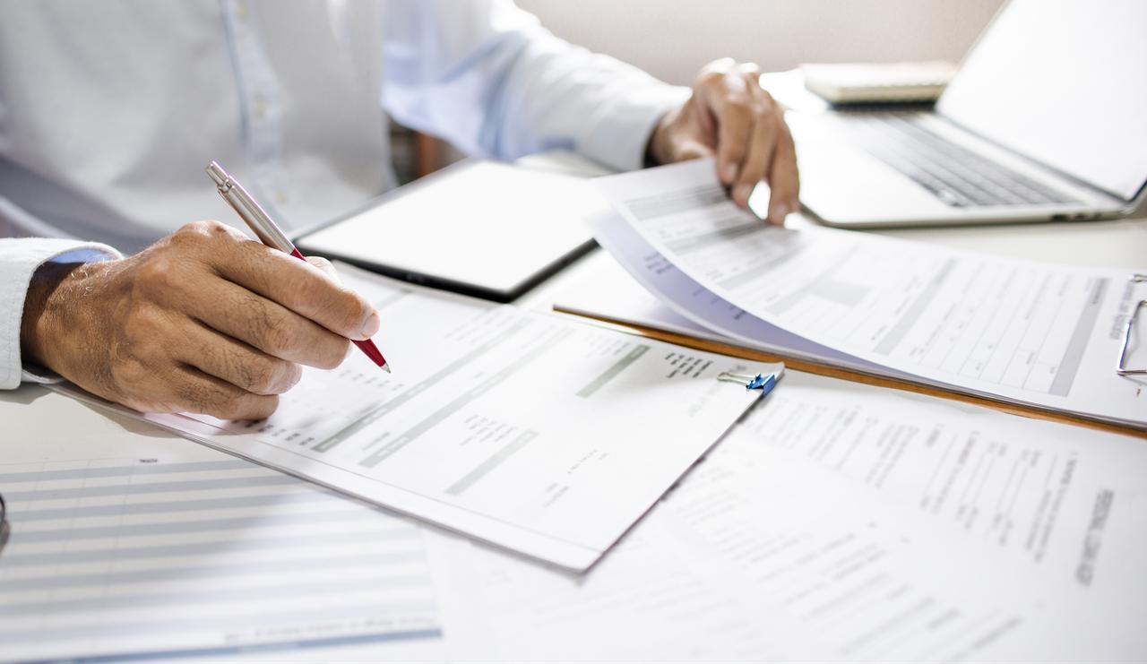 A person reviews paperwork and fills out forms at a desk beside a laptop. (Adobe Stock Photo)