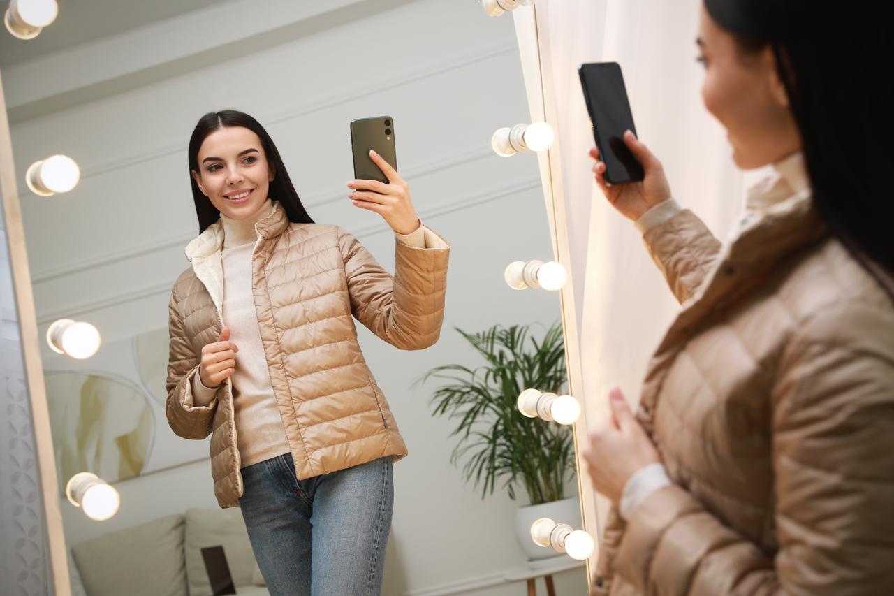 A woman takes a mirror selfie while trying on a jacket, illustrating social media's influence on fashion choices. (Adobe Stock Photo)