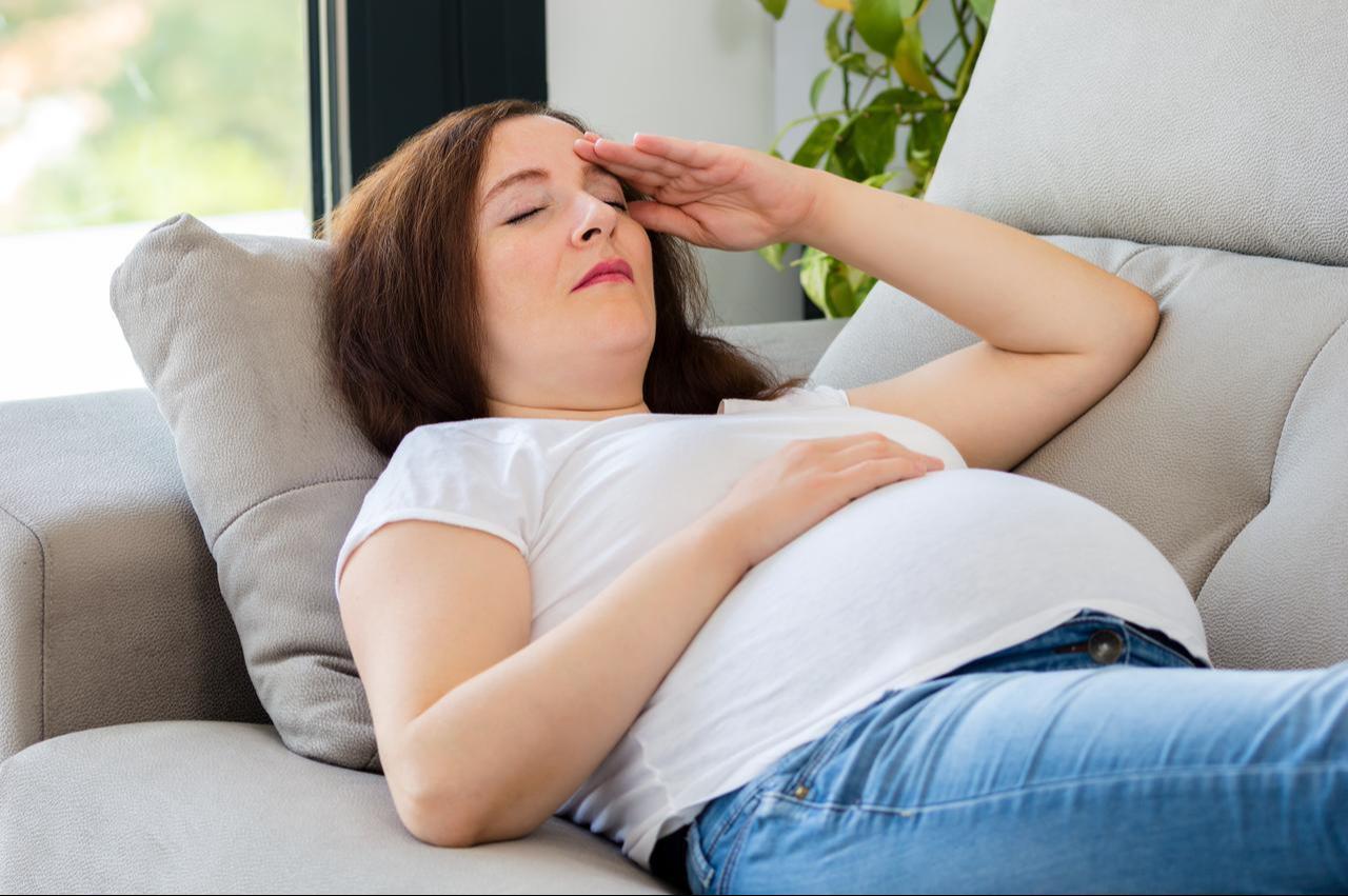 A pregnant woman rests on a sofa while holding her forehead, illustrating fatigue and discomfort during pregnancy. (Adobe Stock Photo)