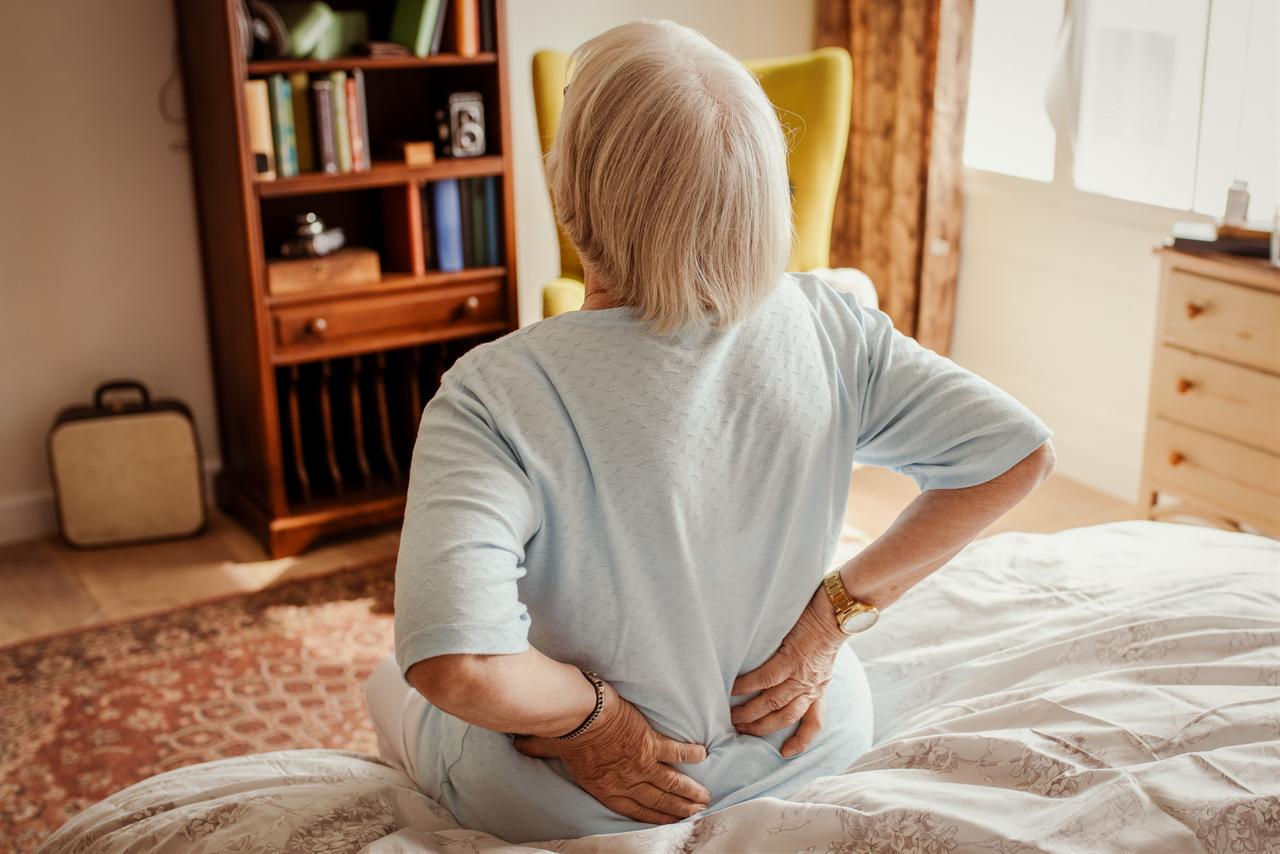 An elderly woman holds her lower back while sitting on the edge of a bed, illustrating back pain among older adults. (Adobe Stock Photo)