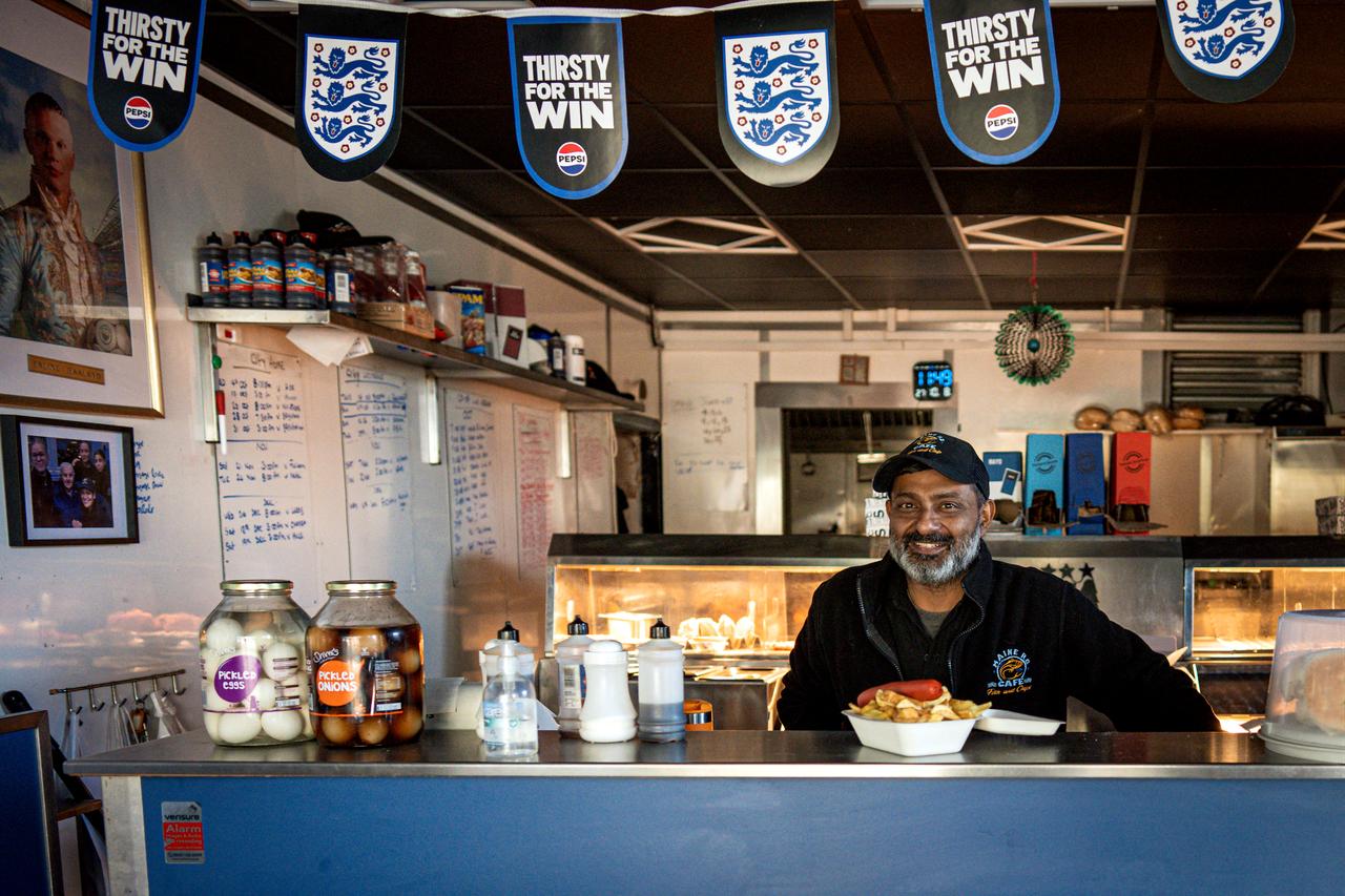 A cafe worker poses behind the counter beneath England football banners at a local business in Manchester, England, Oct. 9, 2026. (AFP Photo)