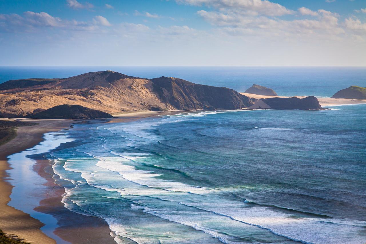 Waves break along a rugged coastal landscape bordered by hills and sandy shoreline. (Adobe Stock Photo)