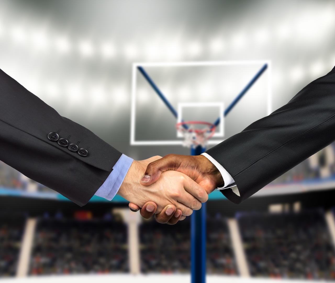 Business representatives shake hands in front of a basketball hoop, illustrating a sports-related partnership or commercial agreement. (Photo via Adobe Stock)