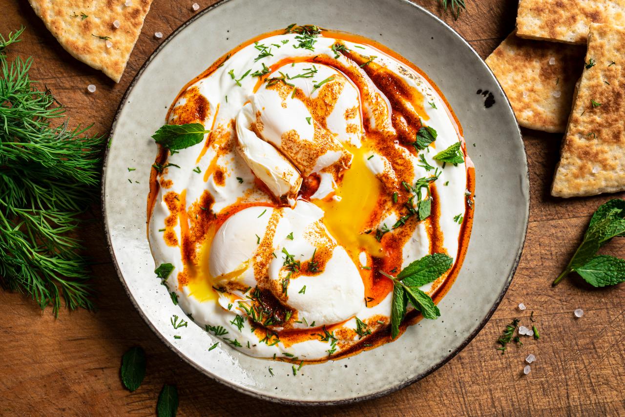 Classic cilbir prepared with poached eggs, thick yogurt, chili-infused oil, and fresh herbs, served with bread on a wooden table setting. (Adobe Stock Photo)
