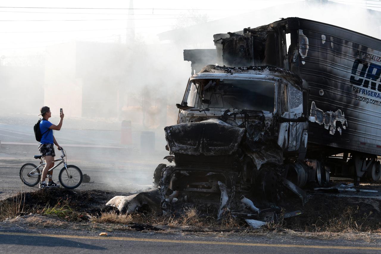 A man riding a bicycle takes a photo of a burned truck, allegedly set on fire by organised crime groups in response to an operation to arrest a high-priority security target, on a highway near Acatlan de Juarez, Jalisco state, Mexico on February 22, 2026. (AFP Photo)