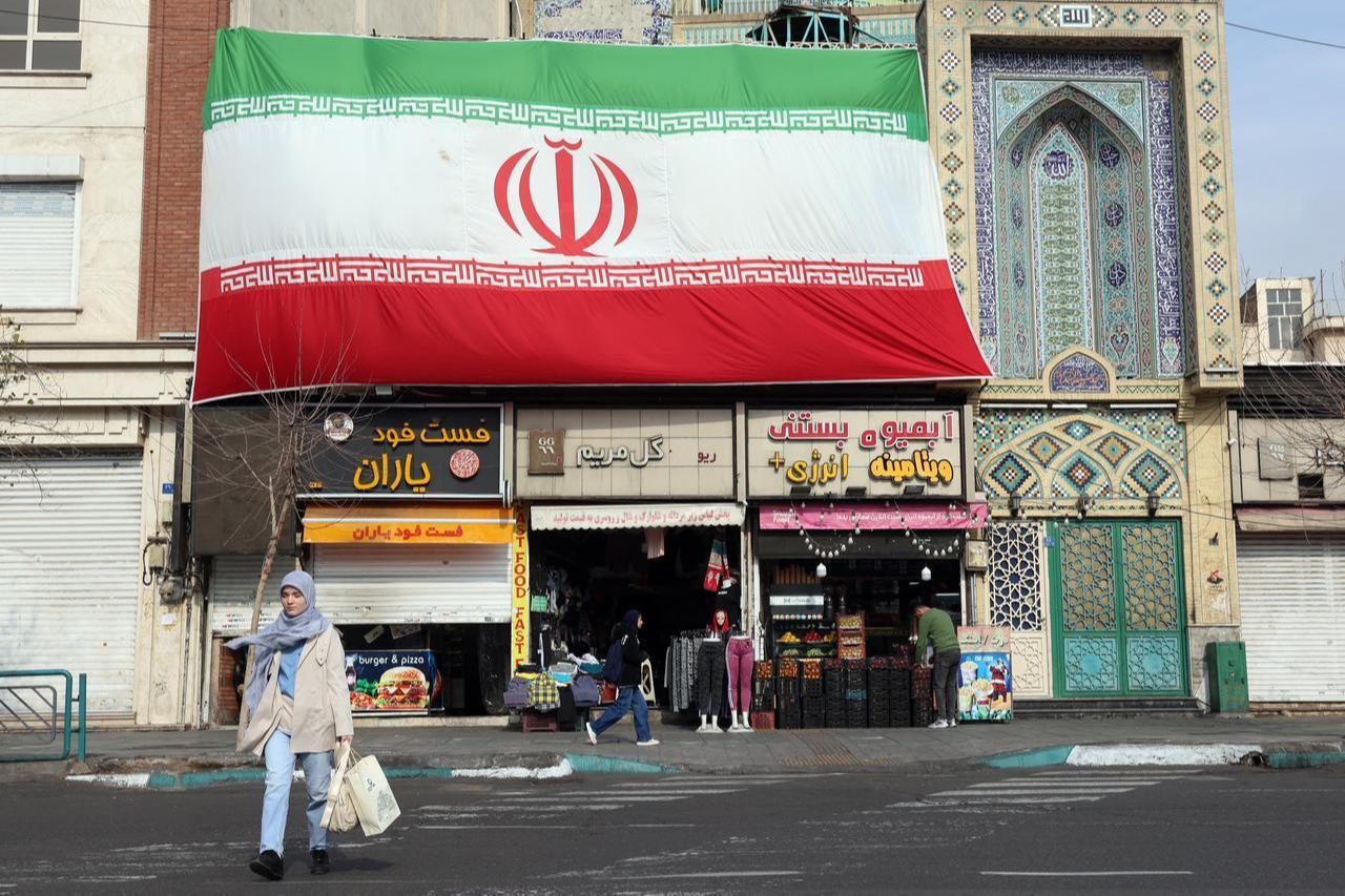 An Iranian woman walks past a huge national flag hanging above shops in Tehran on February 6, 2026. (AFP Photo)