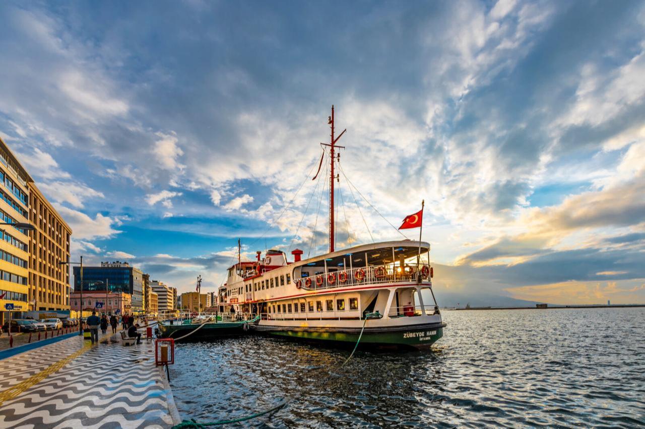 A ferry with the Turkish flag docked at Izmir’s Kordon, with the wave-patterned promenade, cityscape, and a dramatic sunset sky in the background. (Adobe Stock Photo)
