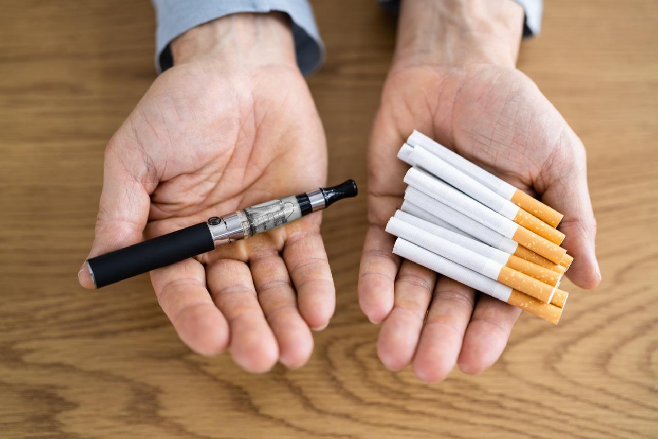 A person holds an e-cigarette in one hand and traditional cigarettes in the other, illustrating comparison, March 2026. (Adobe Stock Photo)