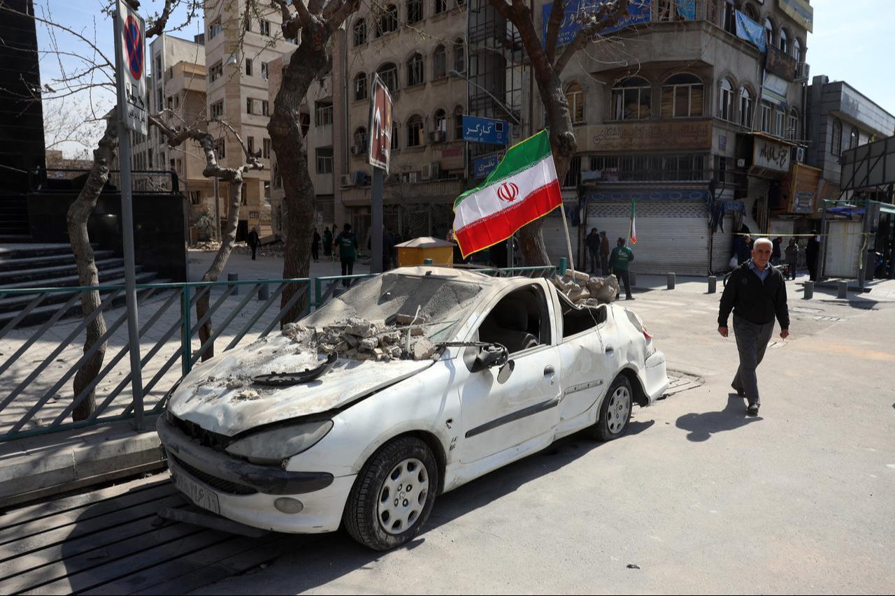 A man walks past an Iranian flag fluttering above the wreckage of a car in central Tehran, on March 4, 2026. (AFP Photo)