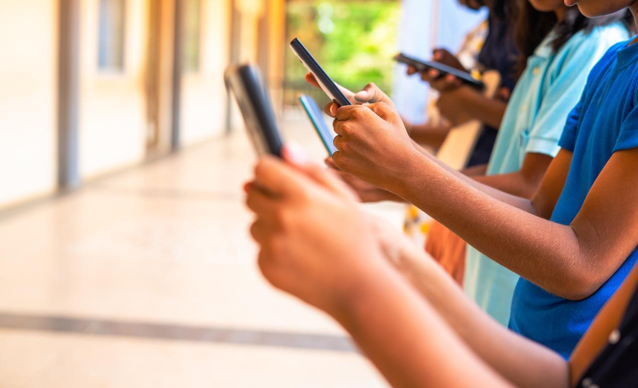 Photo shows a group of children using smartphones in a school corridor, highlighting the role of social media, gaming, technology, and education in their daily lives. (Adobe Stock Photo)