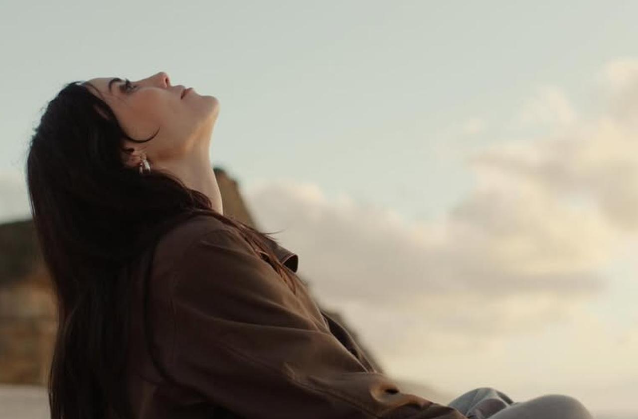 Cansu Dere sits by the coast, looking up toward the sky in a quiet moment captured during filming. (Photo via Instagram/@cansudere)