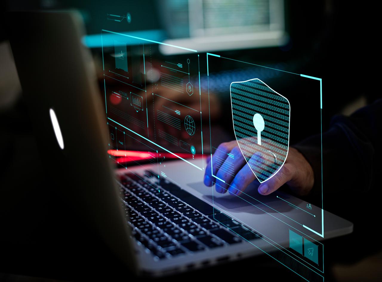 A person uses a laptop with a digital cybersecurity interface and shield icon displayed in an unspecified location, undated. (Adobe Stock Photos)