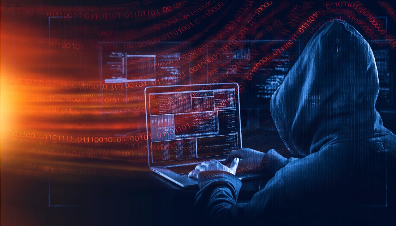 A hooded person uses a laptop with digital code overlays suggesting cyber activity in an unspecified location, undated. (Adobe Stock Photo)