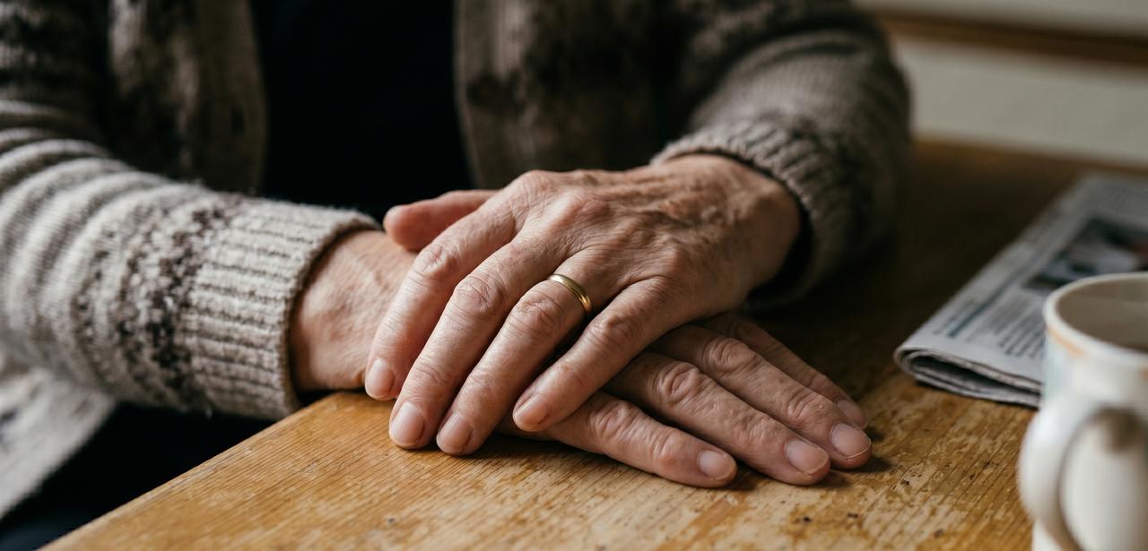 An older woman’s hands rest on a table beside a cup and newspaper in an undated AI-generated image. (Photo generated by Gemini)