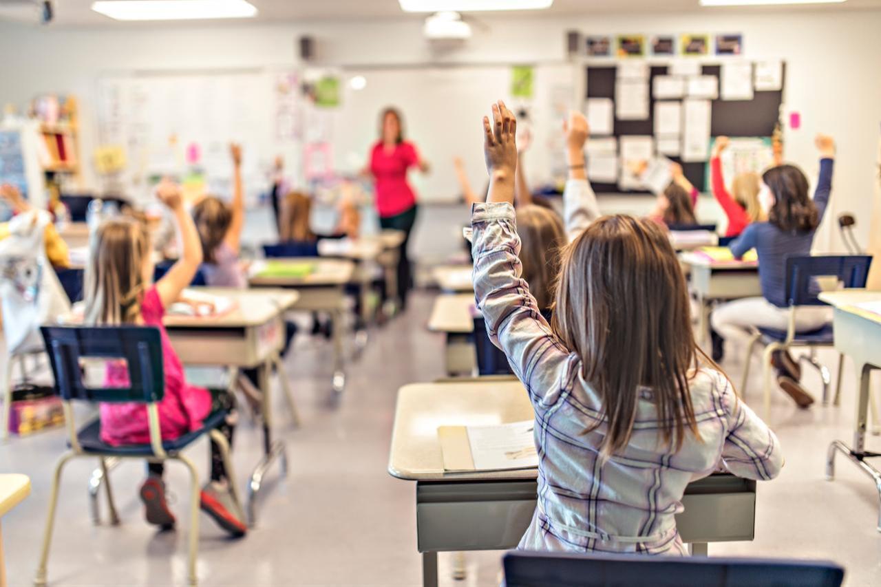 A classroom scene shows students raising their hands to respond to the teacher’s questions. (Adobe Stock Photo)