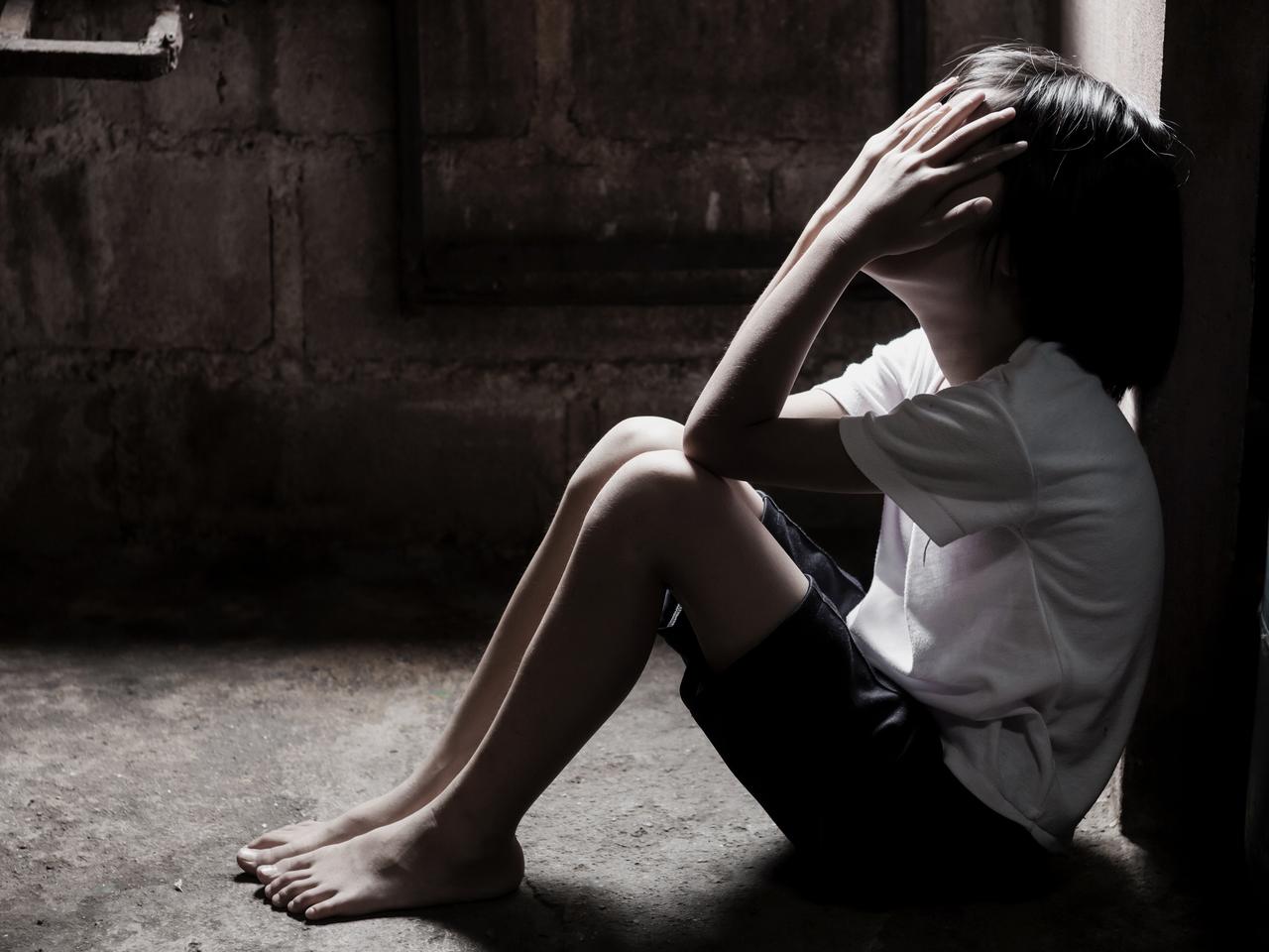 A child sits on the floor with hands over their face in a dim room (Adobe Stock Photo)