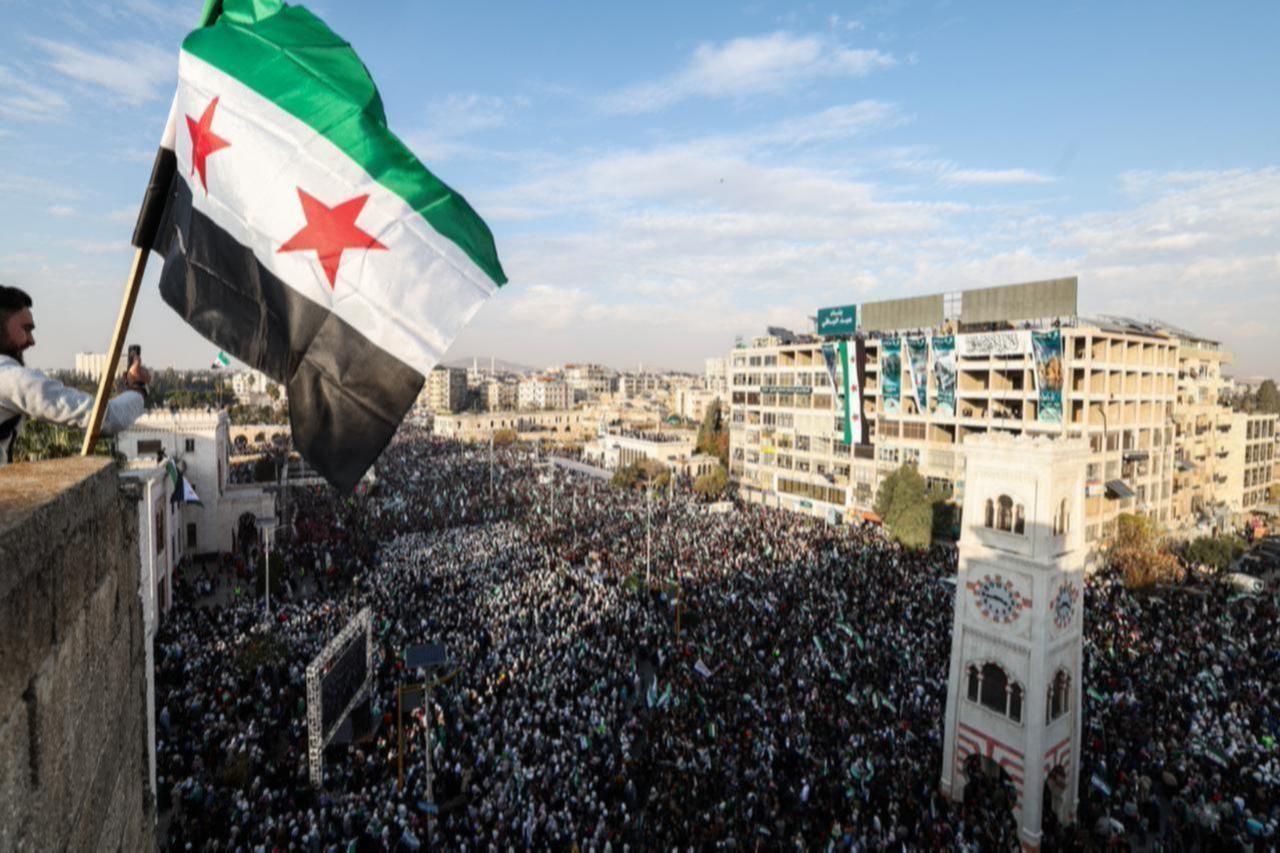 A Syrian flag flutters above crowds gathering during celebrations marking one year since a lightning offensive that eventually toppled the countrys longtime ruler, in central Hama, Syria on December 5, 2025. (AFP Photo)