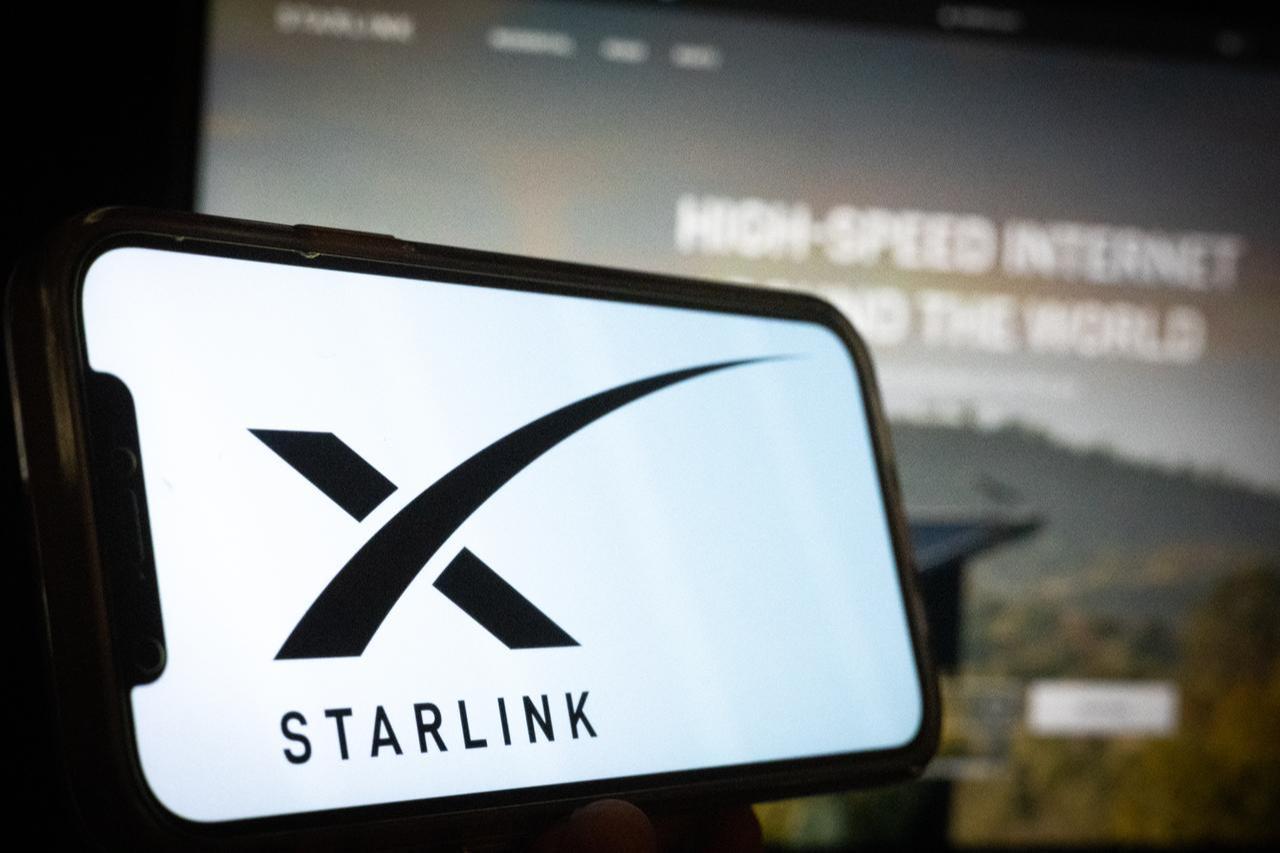 Silhouette of a hand holding a phone with the Starlink logo in front of a satellite internet services website on a computer screen. (Adobe Stock Photo)