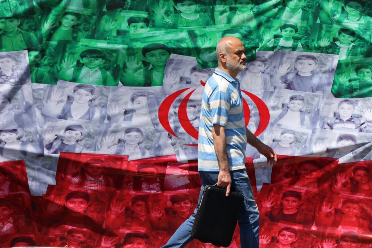 A man walks past an Iranian flag installed along the roadside in Tehran on April 29, 2026, depicting images of children killed on the first day of the war in a US-Israeli missile strike on a school in the southern Iranian city of Minab. (AFP Photo)