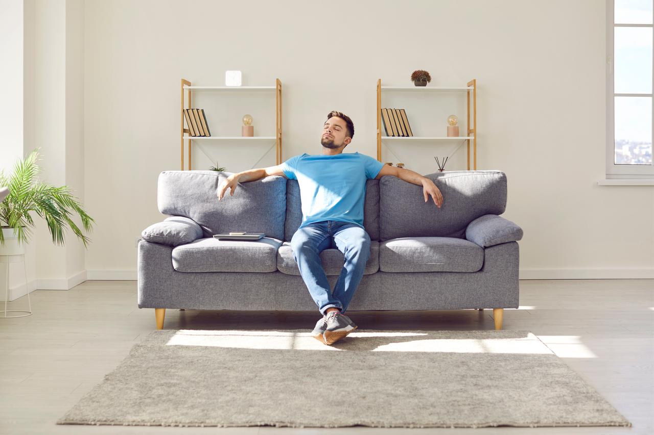 A man relaxes on a sofa in a sunlit living room at home. (Adobe Stock Photo)