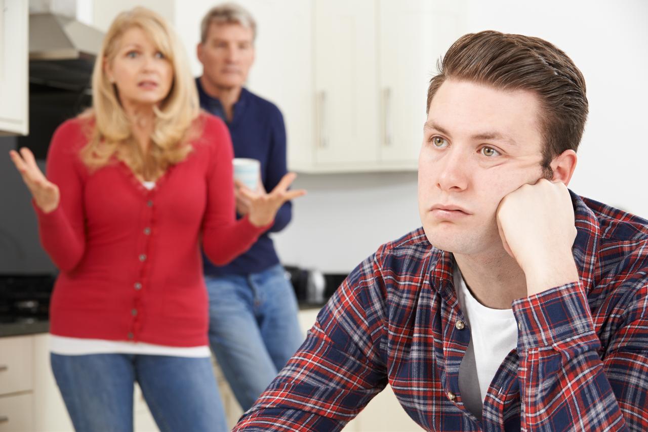 A young man sits apart while two older adults appear to argue in the background inside a home setting. (Adobe Stock Photo)