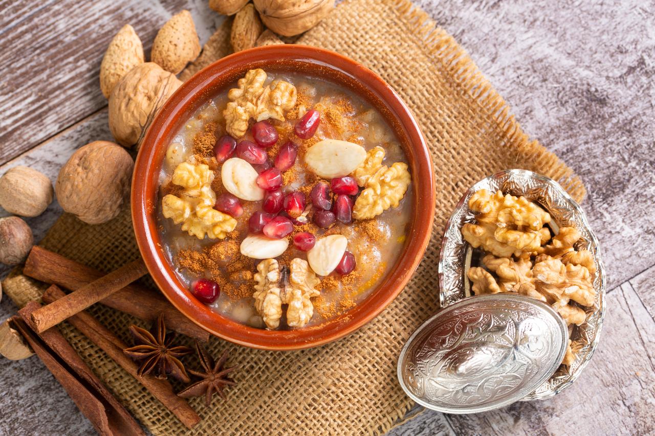 A bowl of asure topped with walnuts, almonds, pomegranate seeds, and cinnamon beside nuts and spices, accessed on May 8, 2026. (Adobe Stock Photo)