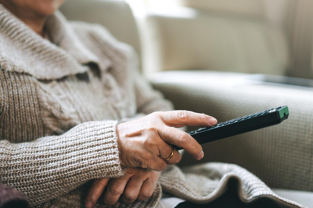 An elderly woman watching television while holding a remote control. (Adobe Stock Photo)