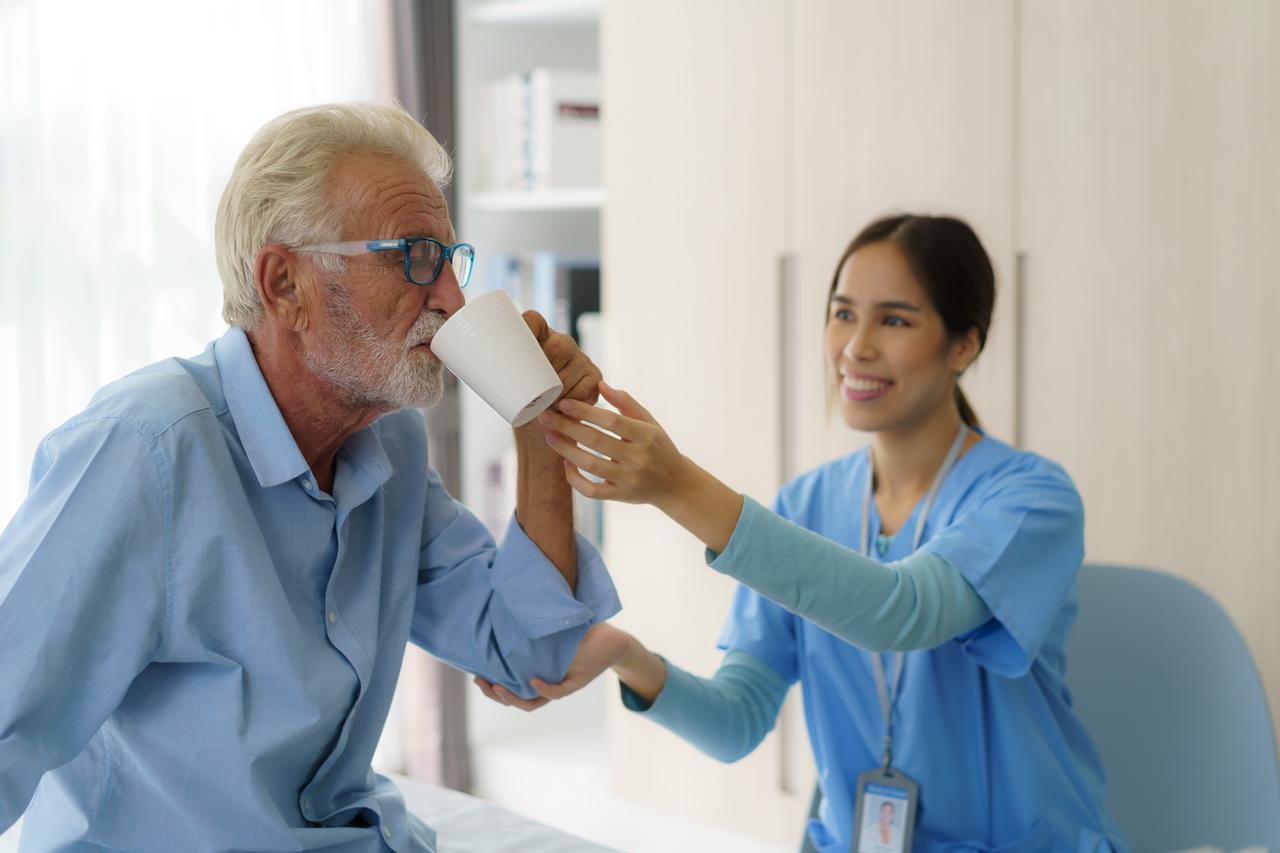 A caregiver helps an older man drink from a cup (Adobe Stock Photo)