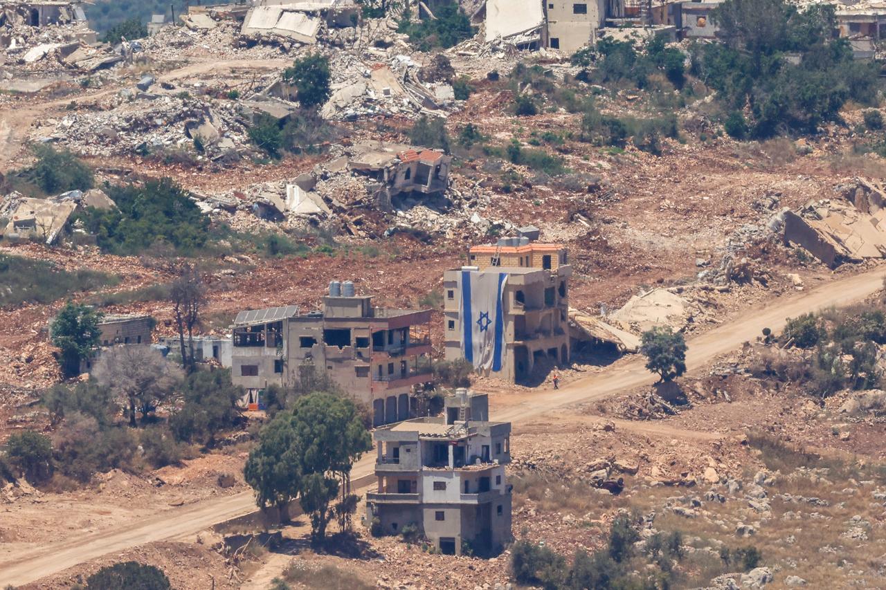 This picture taken from a position in the Upper Galilee in northern Israel, a man walking past an Israeli flag covering part of a destroyed building in the southern Lebanese village of Taybeh near the border on June 18, 2026. (AFP Photo)