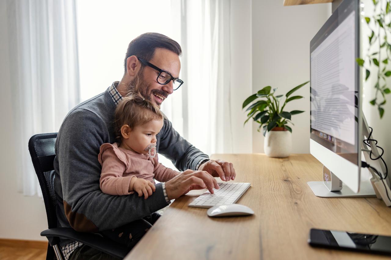 A father works on a computer while holding his child (Adobe Stock Photo)