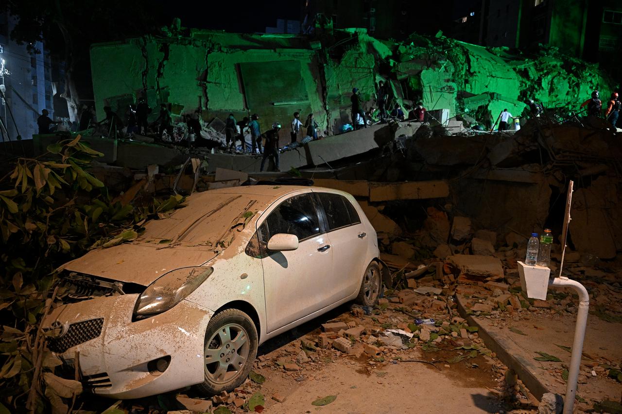 A car crushed by a fallen tree is pictured as people walk in front of a destroyed building following an earthquake in Caracas on June 24, 2026. (AFP Photo)