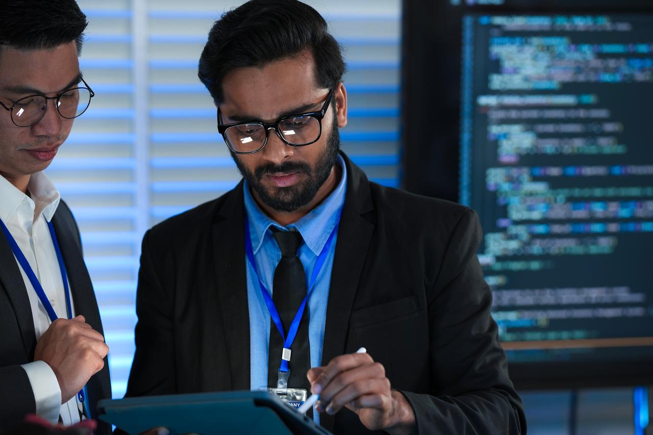 Cybersecurity professionals review data on a tablet in front of a screen displaying computer code. (Adobe Stock Photo)