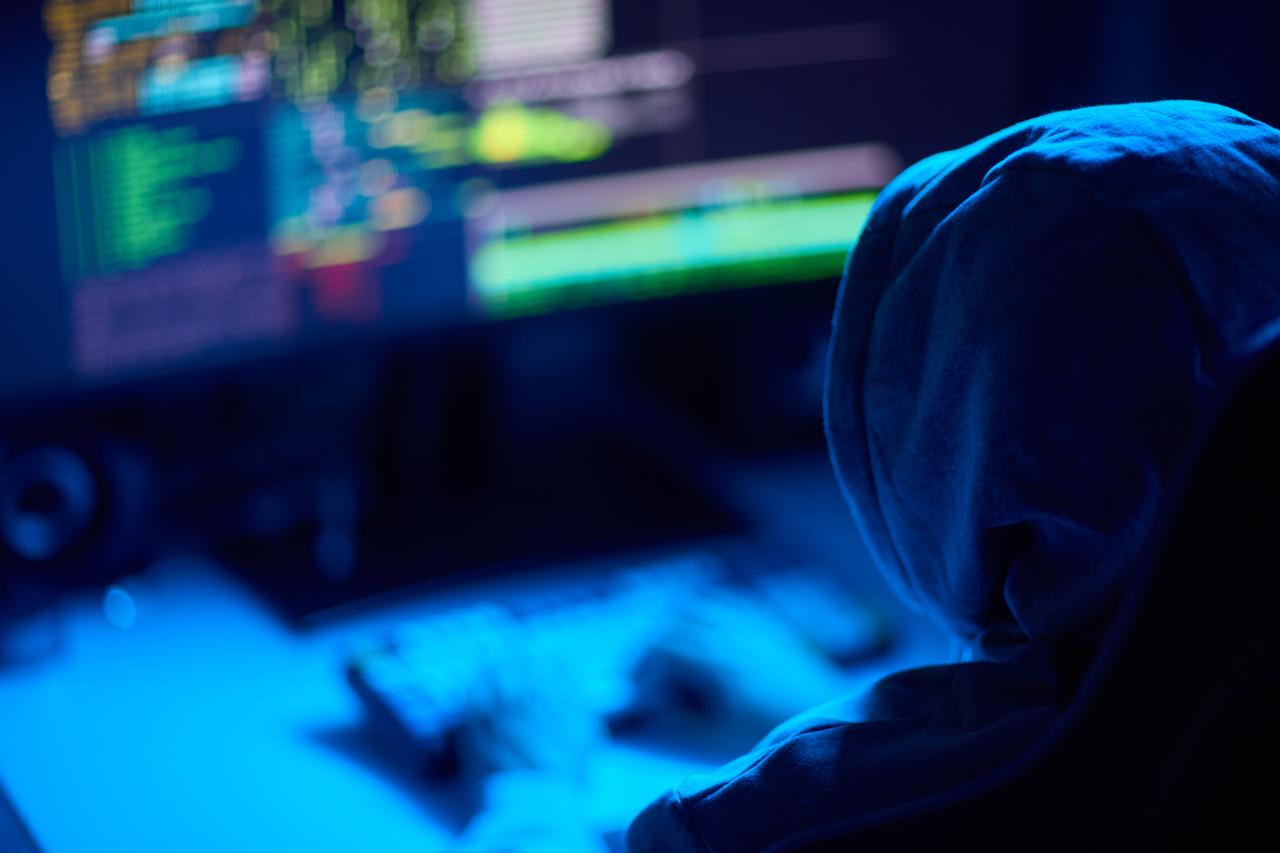 A hooded person uses a computer in front of screens displaying code. (Adobe Stock Photo)