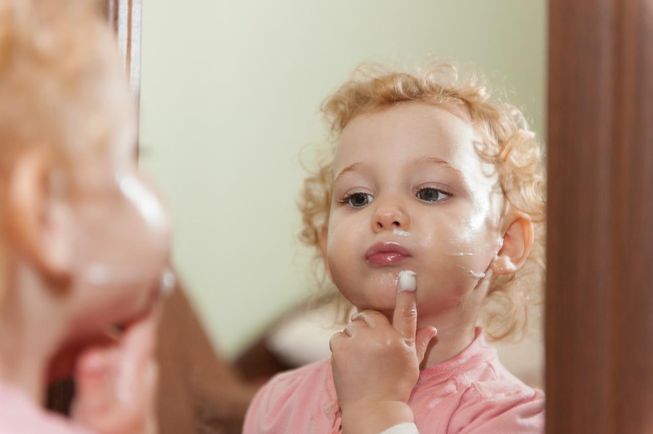 A child applies face cream while looking in a mirror. Adobe Stock Photo)