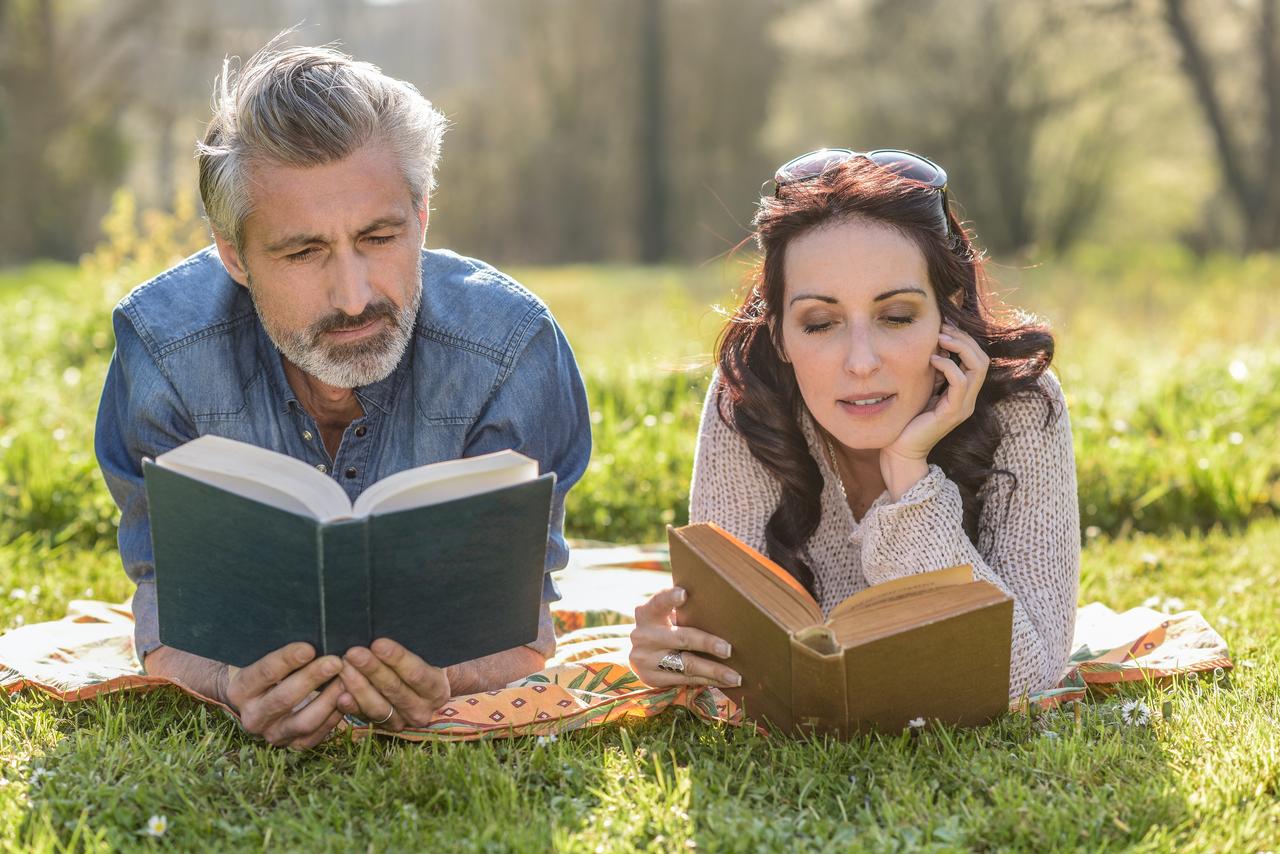 A couple reads books while lying on a blanket in a park. (Adobe Stock Photo)