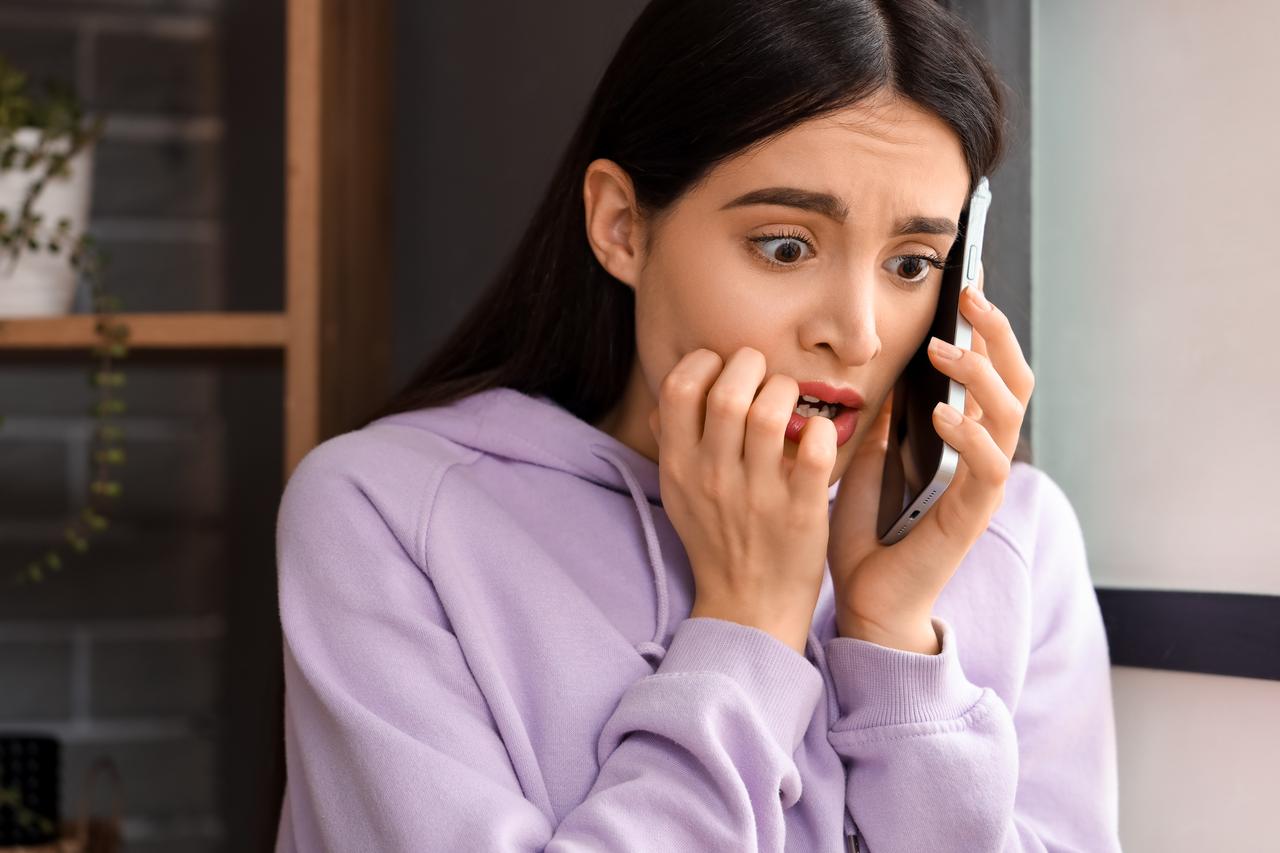 Scared young woman talking by mobile phone at home. (Photo via Adobe Stock)