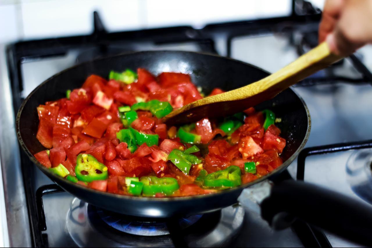 A wooden spoon stirs fresh tomatoes and sliced green peppers in a pan on the stove, accessed on June 3, 2026. (Adobe Stock Photo)