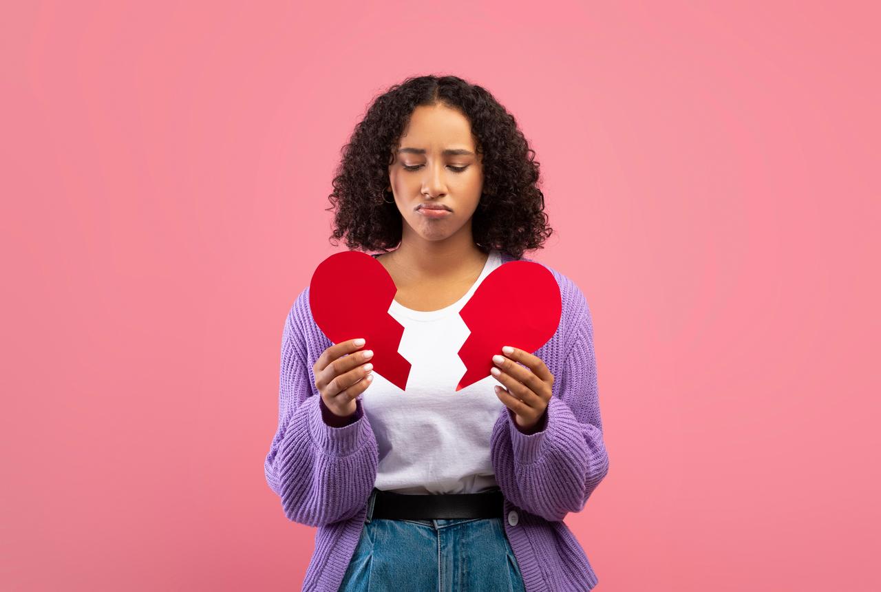 Unhappy youmg woman holding halves of broken heart. (Photo via Adobe Stock)