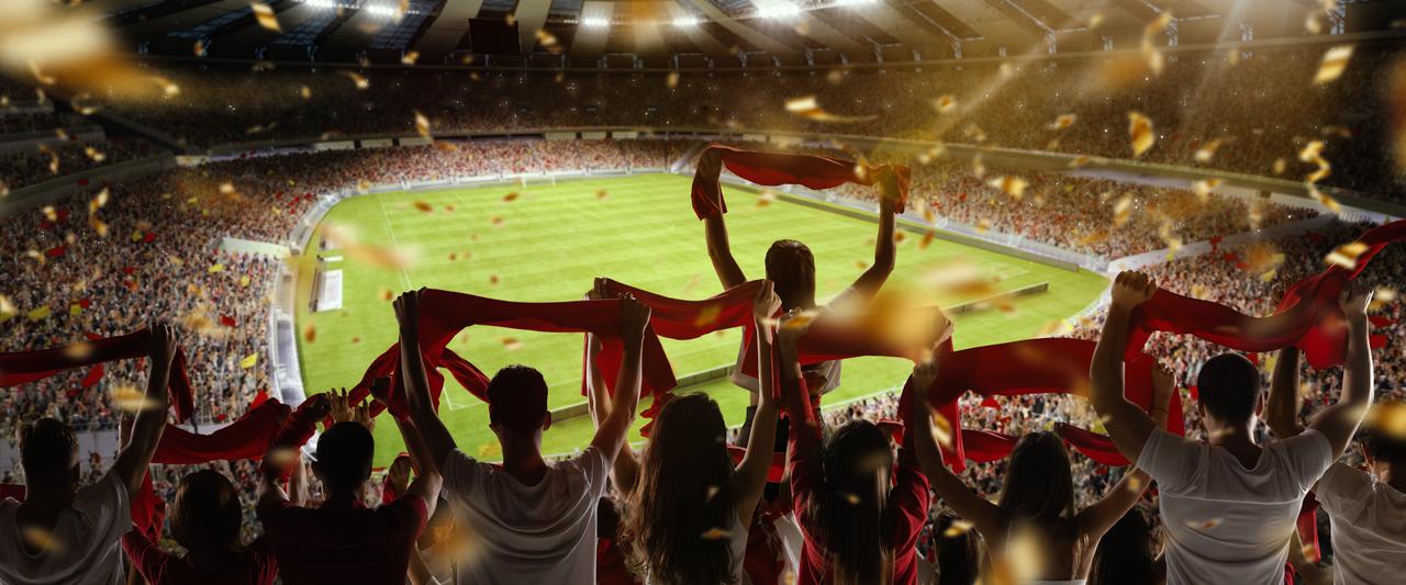 Football fans cheering their team with colorful scarfs at crowded stadium at evening time. (Photo via Adobe Stock)