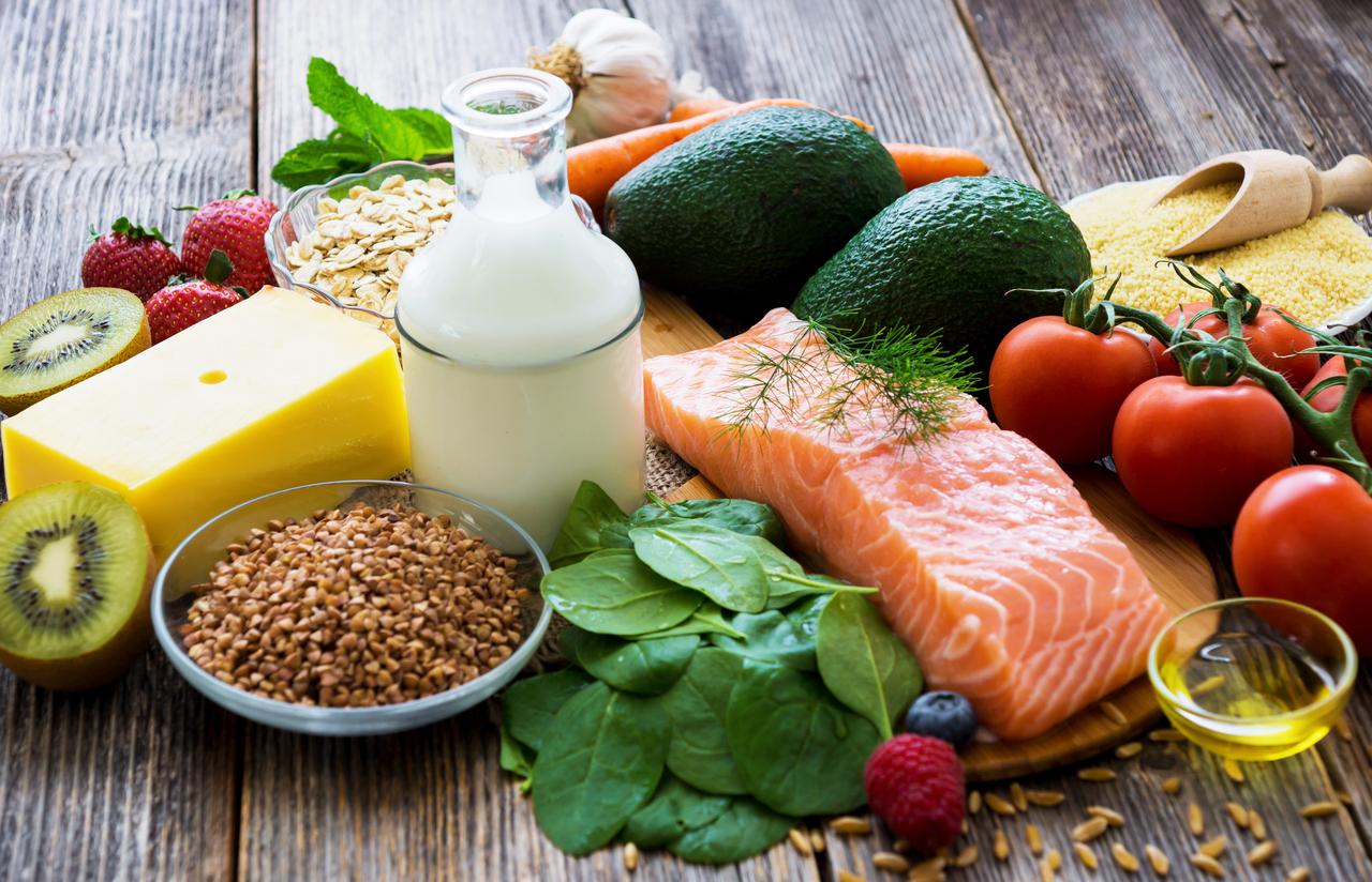Healthy foods, including salmon, milk, cheese, fruits, and vegetables, are displayed on a table. (Adobe Stock Photo)