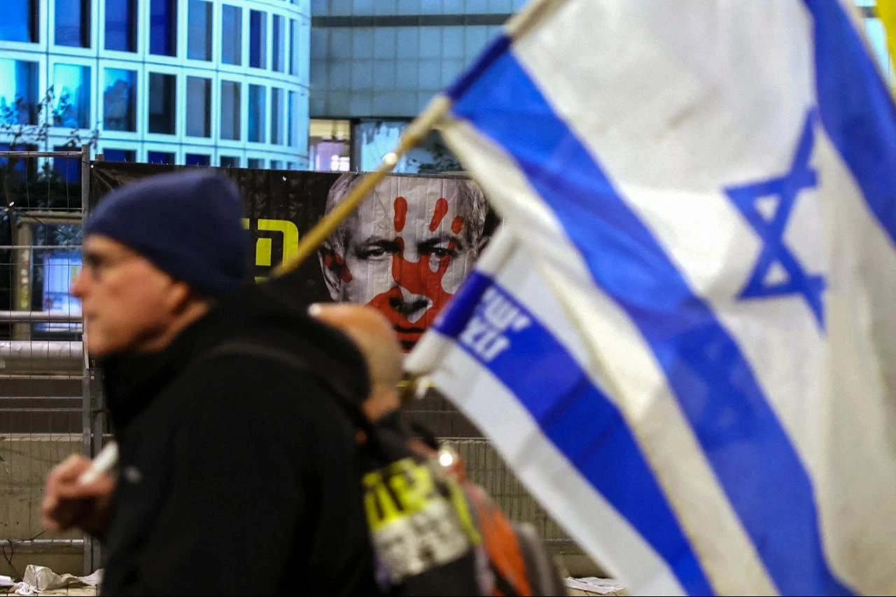 A demonstrator carrying an Israeli flag walks past a banner bearing the portrait of Israeli Prime Minister Benjamin Netanyahu during a protest in front of the Israeli Defence Ministry in Tel Aviv, January 25, 2025. (AFP Photo)