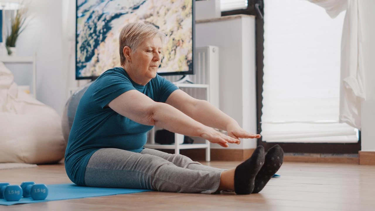 An older woman performs a seated stretching exercise on a mat at home. (Adobe Stock Photo)