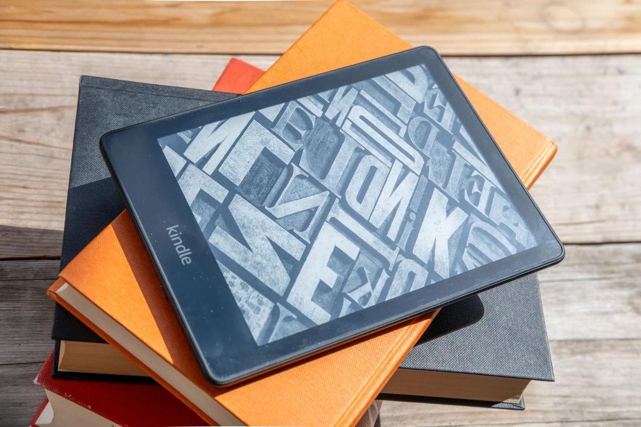An Amazon Kindle rests on a stack of printed books on a wooden surface. (Adobe Stock Photo)