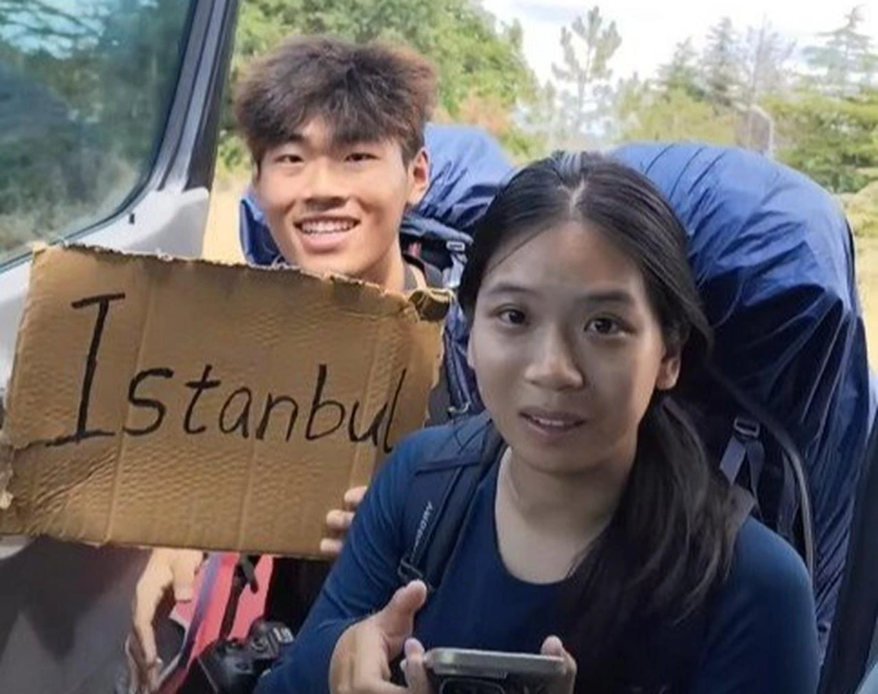 Two Chinese travelers carrying large backpacks hold up a cardboard sign marked “Istanbul” while hitchhiking in Türkiye. (Photo via Türkiye Daily)