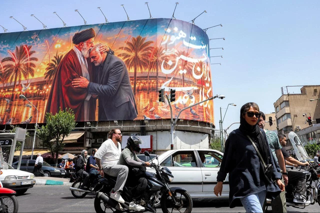 People cross a street past a billboard depicting Iran's slain supreme leader Ali Khamenei kissing the slain Islamic revolutionary guards commander Qasem Soleimani near the shrine of Imam Hussein Iraq's holy city of Karbala, displayed on the facade of a building in Tehran, June 29, 2026. (AFP Photo)