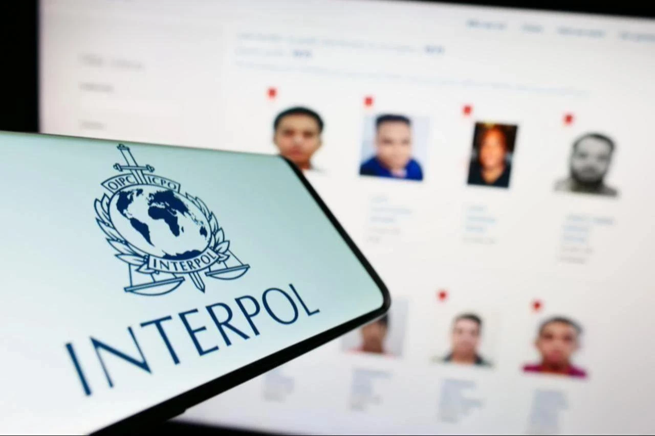 Interpol logo displayed on a mobile phone in front of a screen showing wanted suspects, accessed on June 14 2026. (Adobe Stock Photo)