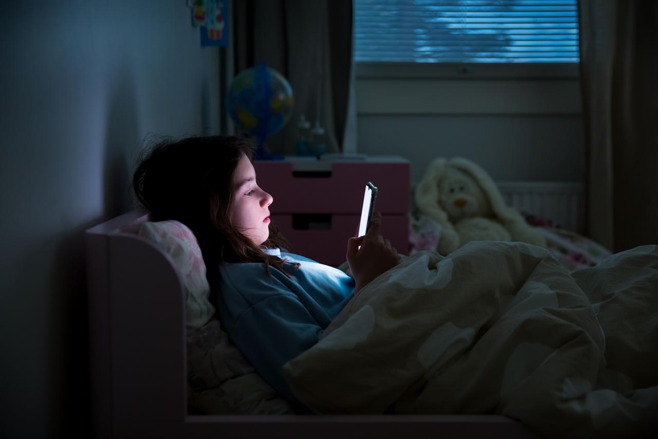 A child using smart phone lying in bed late at night, scrolling. (Photo via Adobe Stock)