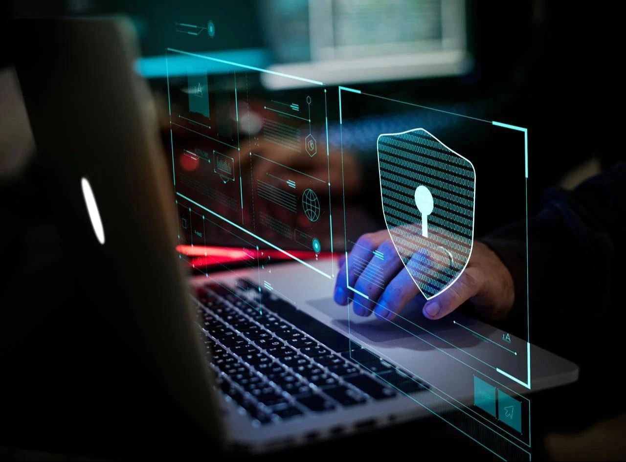 A person uses a laptop with a digital cybersecurity interface and shield icon displayed in an unspecified location, undated. (Adobe Stock Photo)